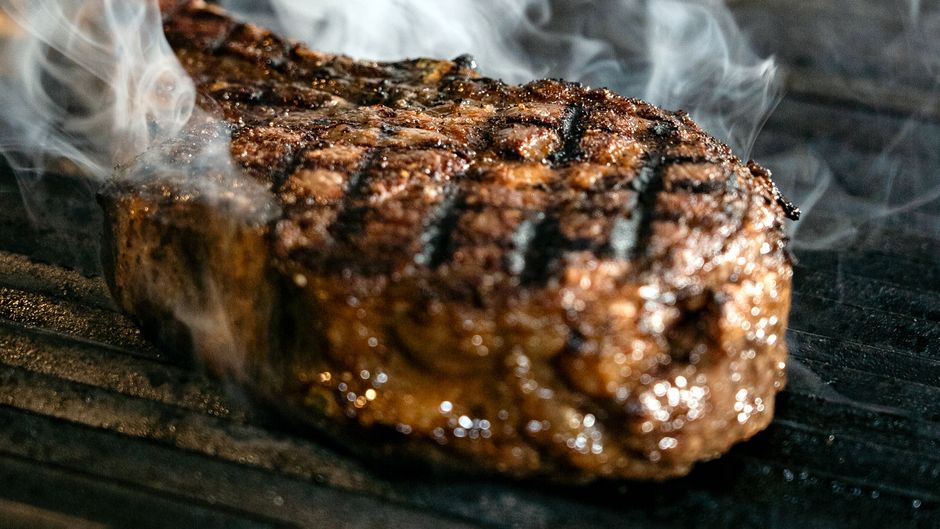 Close-up of a grilled steak with smoke rising, highlighting its juicy texture and grill marks.